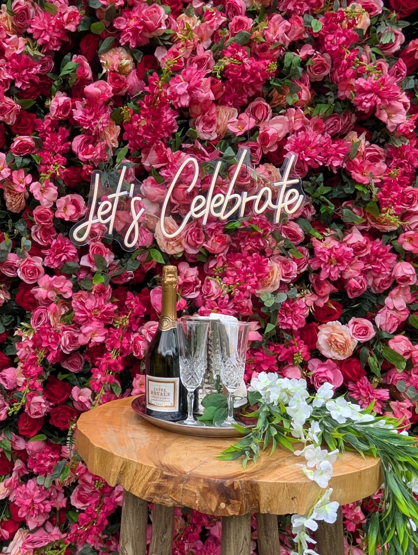 Edith - Detail A champagne bucket on a wooden table in front of a pink floral backdrop with "Let's Celebrate" sign.