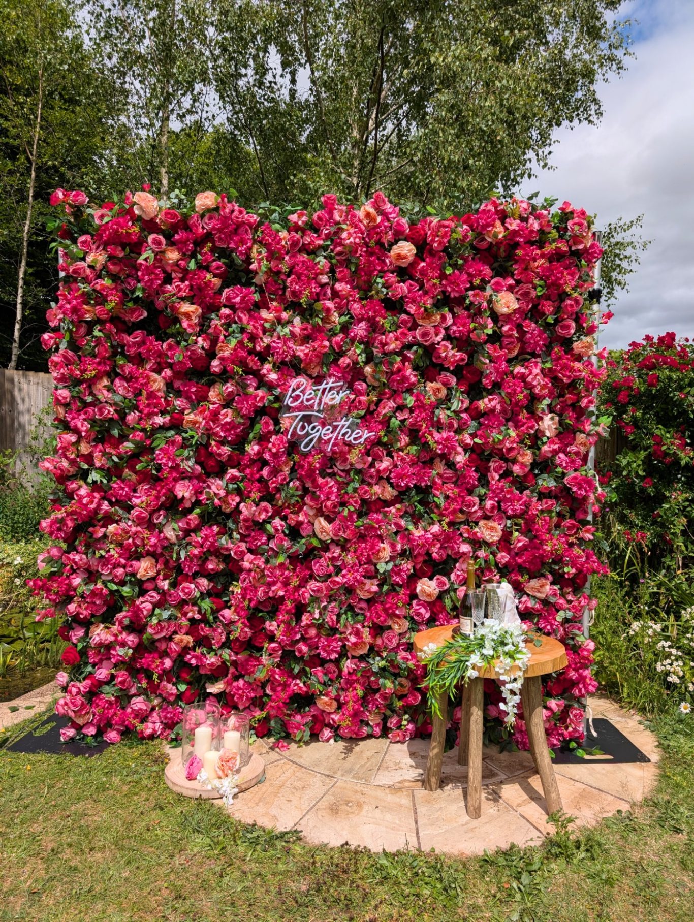 Edith - Size: Regular Wall of vibrant pink flowers with a rustic wooden table in front, set in a lush garden.