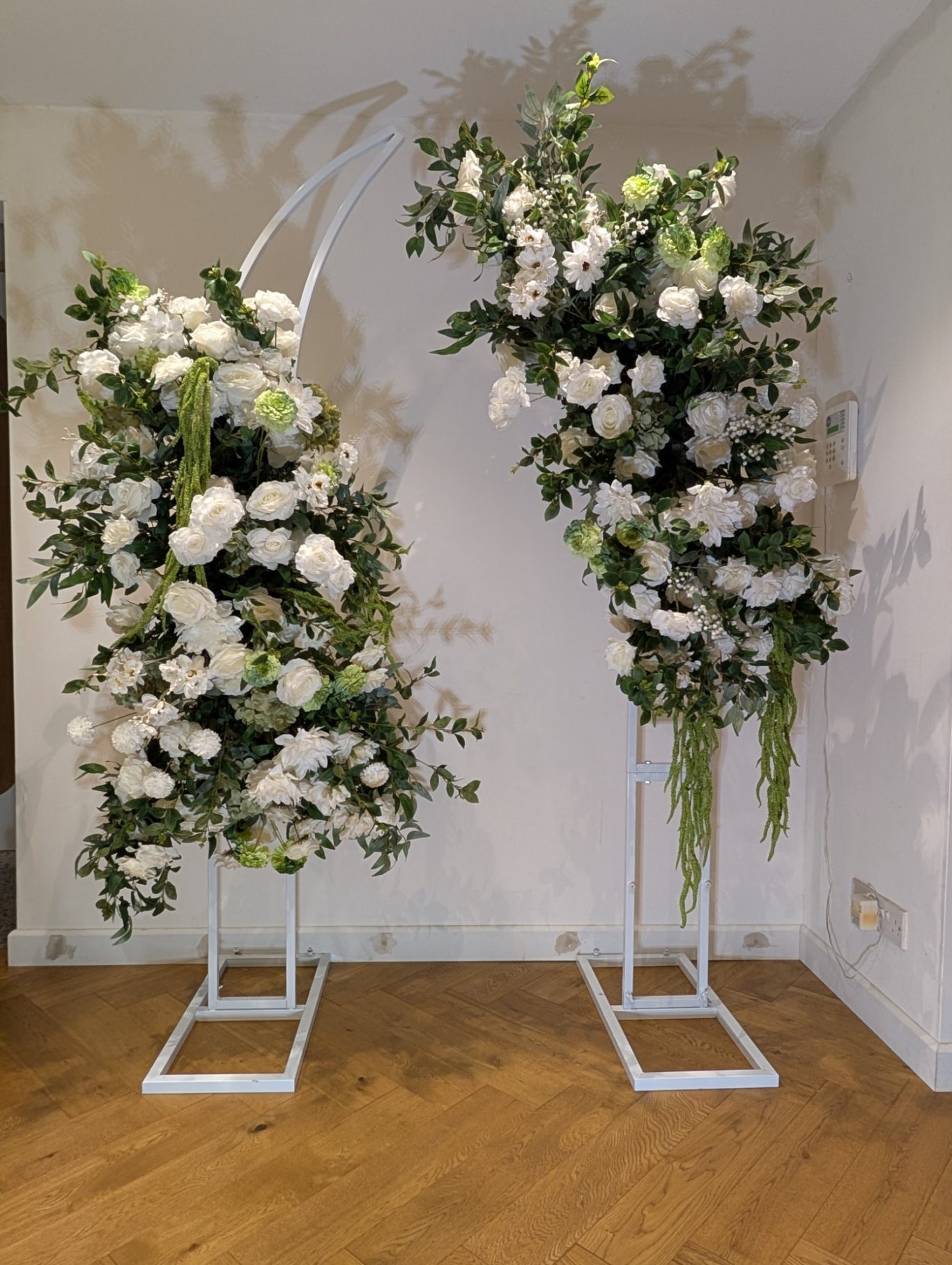 Grace - Broken Arch Two tall floral arrangements with white roses and greenery on white stands, against a light wall.
