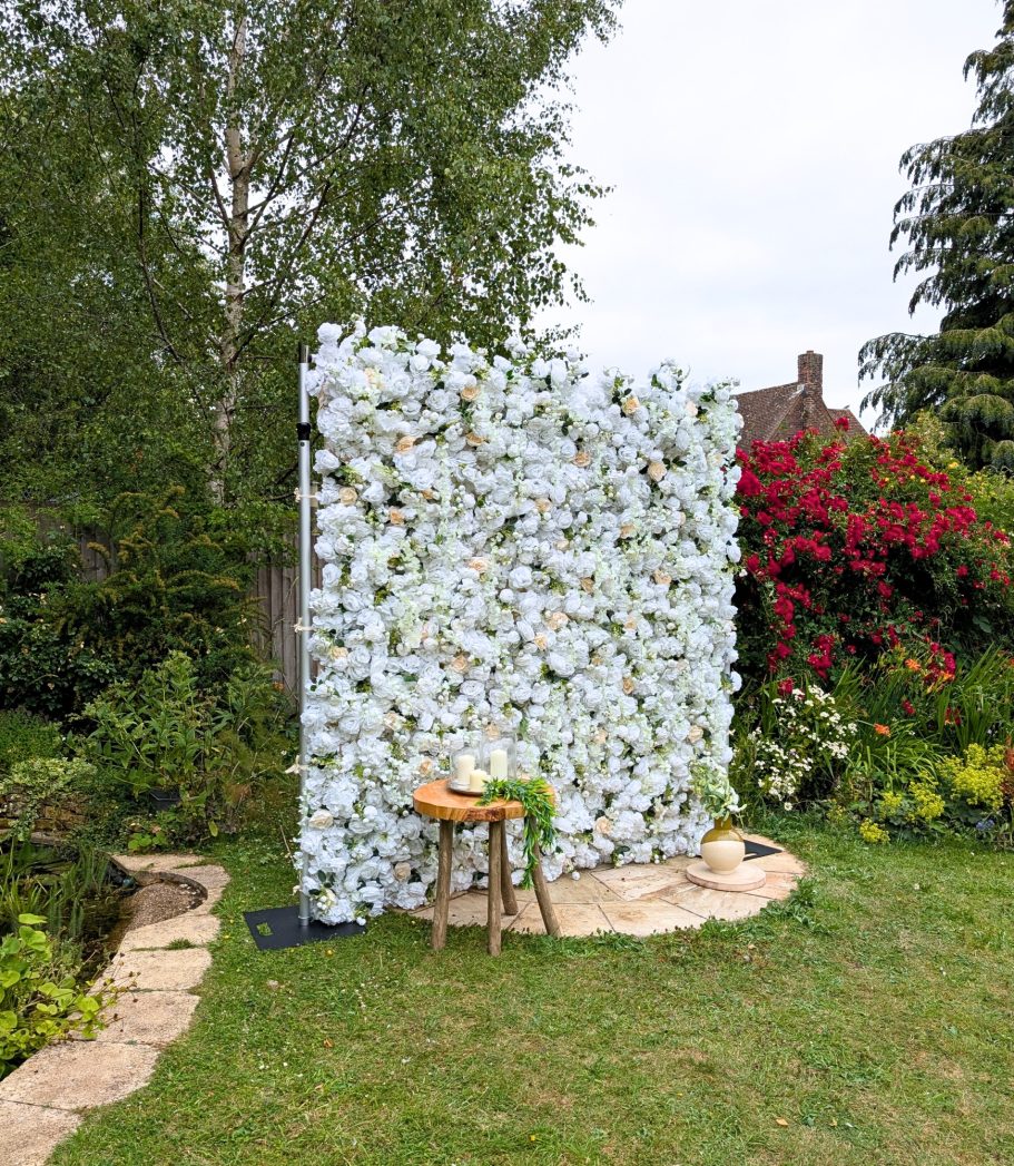 Joy Flower Wall A flower wall backdrop with a small wooden stool in a garden setting.
