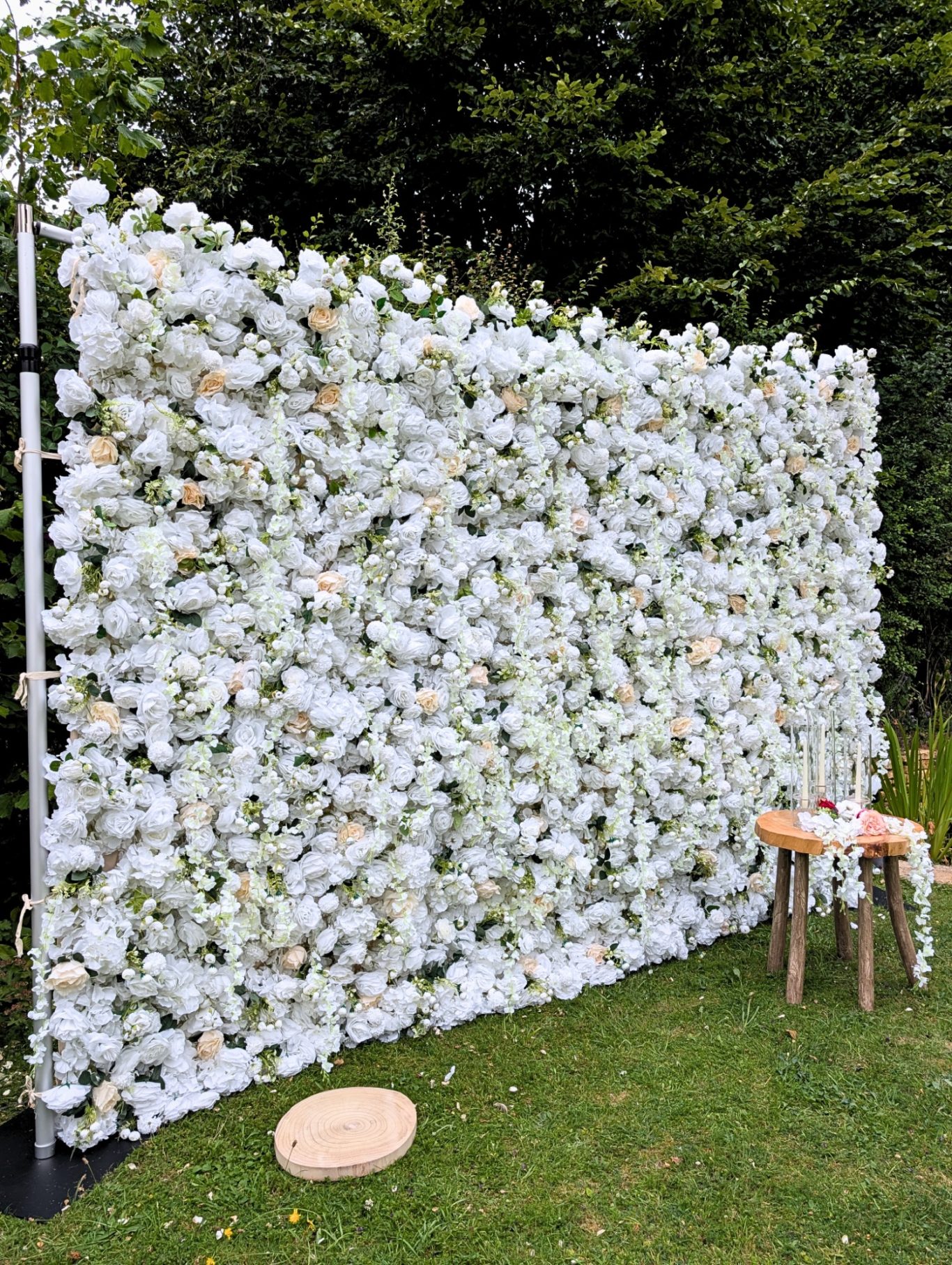 Joy - Size: Large A flower wall display with white blooms and greenery, set in a garden. Two wooden stools nearby.